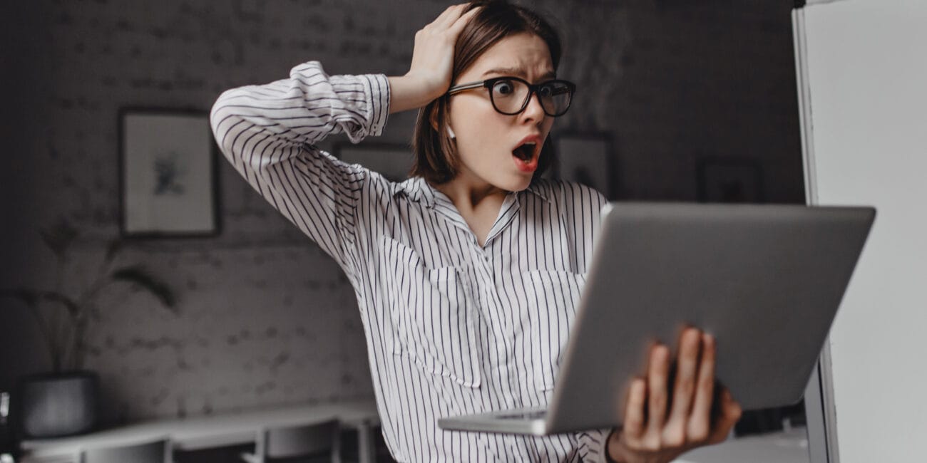 Employee in white shirt and glasses in horror grabs her head because of reports she saw in laptop. Portrait of shocked woman on white office background